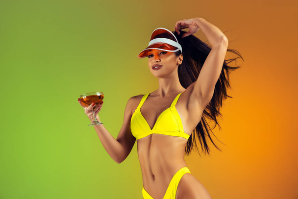 Fashion portrait of young fit and sportive woman with cocktail in stylish yellow luxury swimwear on gradient background. Perfect body ready for summertime. - Fotografie, Obrázek