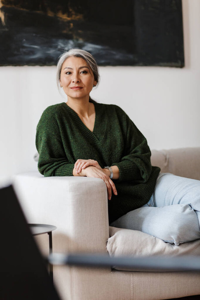 Photo of gray-haired attractive woman looking at camera while sitting on couch in living room - Fotó, kép