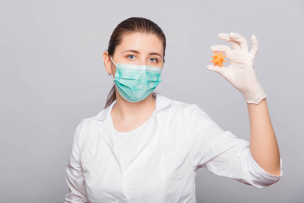 Doctor woman in a white coat, mask and gloves holds a mock virus in her hands. Studio photo on a white background. - Foto, immagini
