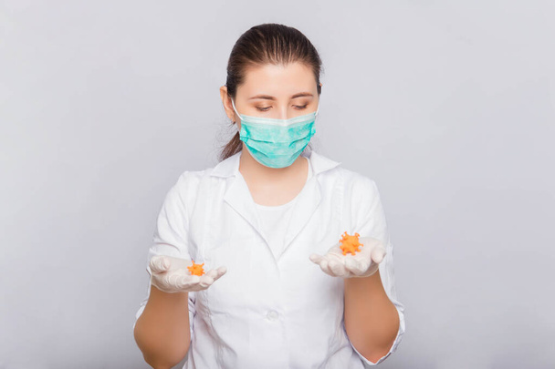 Dokter vrouw in een witte jas, masker en handschoenen houdt een mock virus in haar handen. Studio foto op een witte achtergrond. - Foto, afbeelding