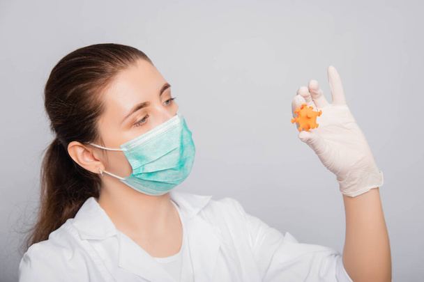 Doctor woman in a white coat, mask and gloves holds a mock virus in her hands. Studio photo on a white background. - Foto, Imagen