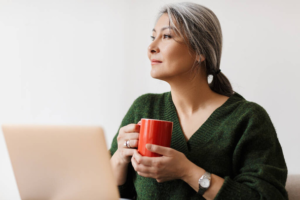 Image of beautiful adult asian woman using laptop computer and drinking coffee while sitting on sofa at home - Foto, afbeelding