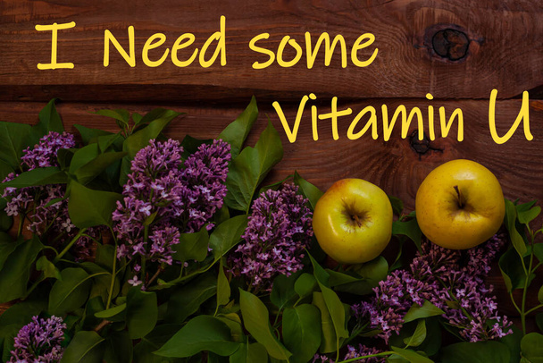 I need some Vitamin U text. Bright yellow apples, purple lilac flowers with fresh green leaves on wooden background flat lay. Colorful Love spring summer fruit backdrop. Cotrasting creative banner. - Φωτογραφία, εικόνα