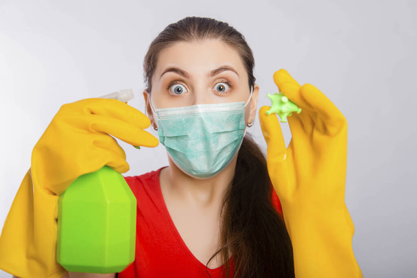 A young woman in a mask and gloves holds a spray bottle and a rag with a model of coronavirus. Concept art for cleaning the premises and the prevention of viral diseases. - Фото, изображение