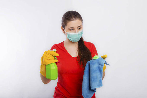 A young woman in a mask and gloves holds a spray bottle and a rag with a model of coronavirus. Concept art for cleaning the premises and the prevention of viral diseases. - Photo, image