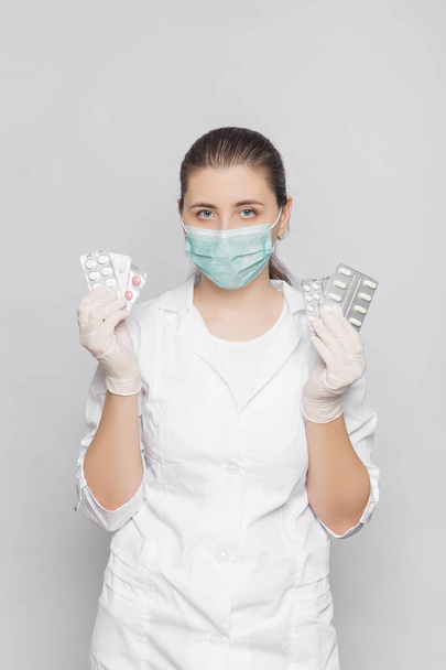 Young woman doctor in a white coat, gloves and a mask holds pills in his hands, standing on a white background. - Fotografie, Obrázek