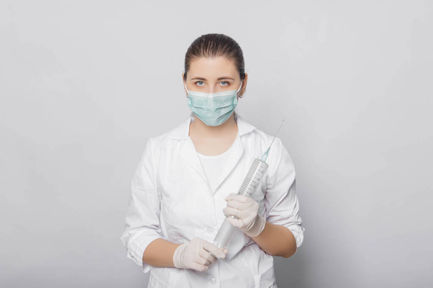 Een vrouw in een witte jas, masker en handschoenen houdt een enorme spuit in haar handen. Portret van een arts in uniform op een witte achtergrond, klaar voor injectie. - Foto, afbeelding