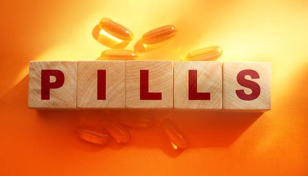 Pills word on wooden cubes with oil capsuless around on orange background. Medical healthcare concept. - Photo, image