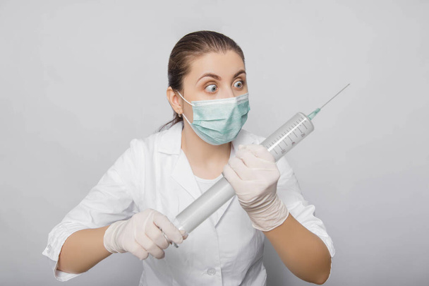 A woman in a white coat, mask and gloves holds a huge syringe in her hands. Portrait of a doctor in uniform on a white background, ready to make an injection. - Foto, imagen