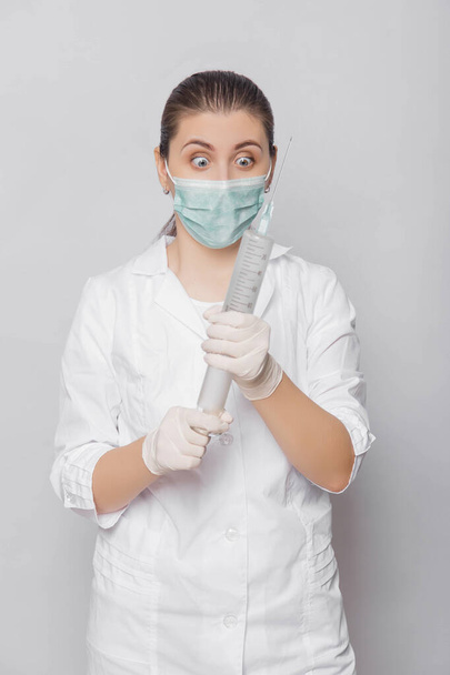 A woman in a white coat, mask and gloves holds a huge syringe in her hands. Portrait of a doctor in uniform on a white background, ready to make an injection. - Foto, Bild