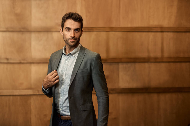 Portrait of a stylish man adjusting the lapel of his blazer while standing alone in a trendy lounge in the evening - Foto, Imagen