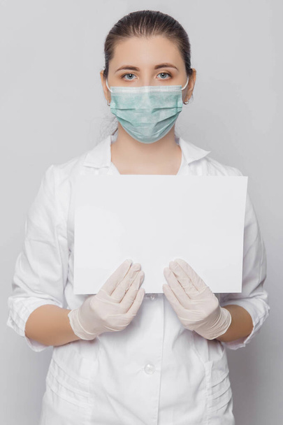 A female doctor in a white coat, mask and gloves holds in her hands an empty sheet of white paper with a place for inscription. Isolated on a white background. - Фото, изображение