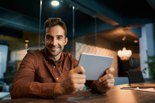 Ritratto di un uomo d'affari sorridente seduto dopo ore da solo alla scrivania del suo ufficio buio e che lavora online con un tablet digitale
 - Foto, immagini