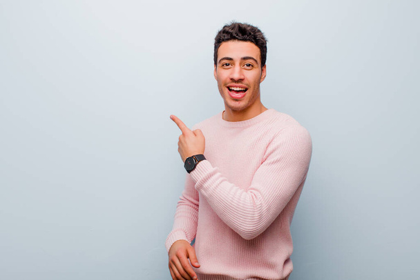 young arabian man looking excited and surprised pointing to the side and upwards to copy space against gray wall - Foto, Bild