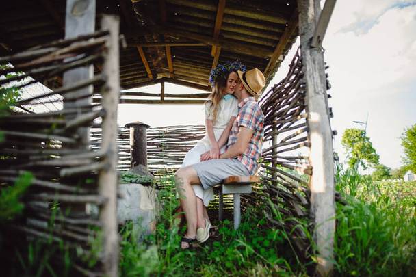 Beautiful girl in a white dress and in a wreath of wild flowers sits in the hands of her boyfriend. Enamored family couple embrace in a rural wooden gazebo. Love story - Photo, image