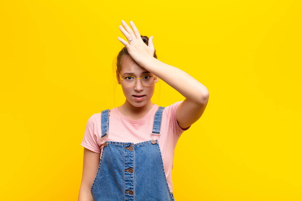 young blonde pretty girl raising palm to forehead thinking oops, after making a stupid mistake or remembering, feeling dumb against yellow wall - Fotoğraf, Görsel