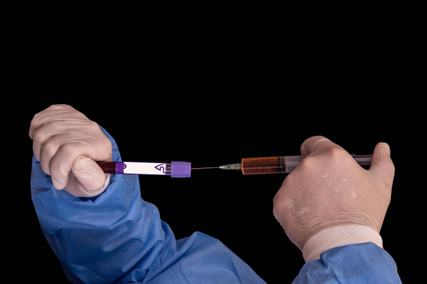 Physician is doing an experiment to create vaccine against the Coronavirus, insert serum from the syringe into a test tube with Covid-19 infected blood, on black background - Photo, Image
