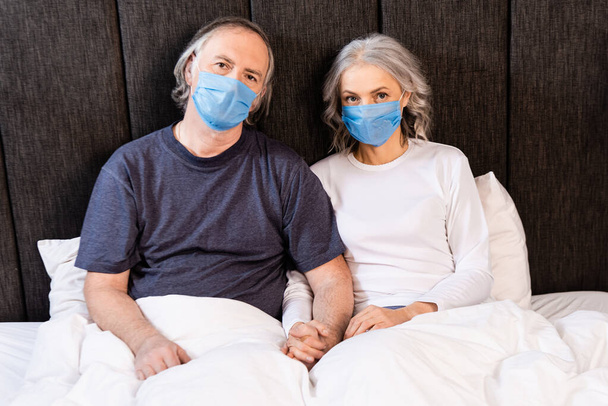 couple d'âge mûr dans des masques médicaux tenant la main au lit
  - Photo, image