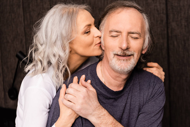 mature woman kissing cheek of happy husband with closed eyes  - Photo, Image