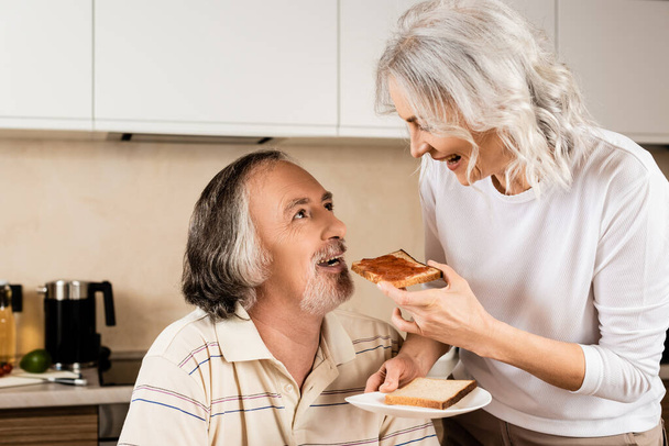 gelukkig volwassen vrouw voeden echtgenoot met zoet toast brood  - Foto, afbeelding