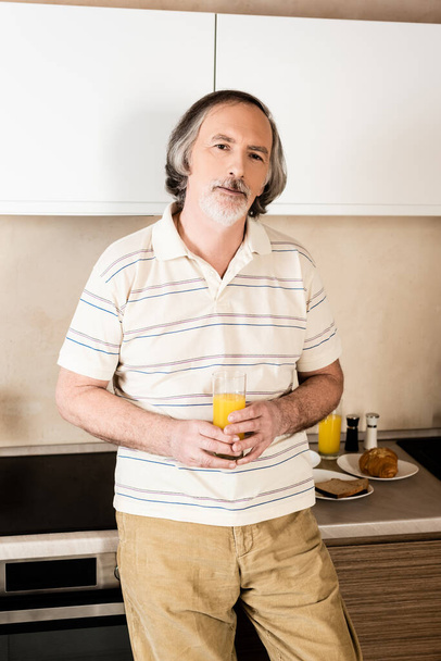 ώριμος και γενειοφόρος άντρας που κρατάει ένα ποτήρι χυμό πορτοκαλιού  - Φωτογραφία, εικόνα