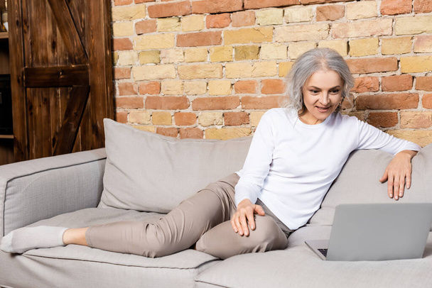 vrolijk en volwassen vrouw kijken film op laptop in woonkamer  - Foto, afbeelding
