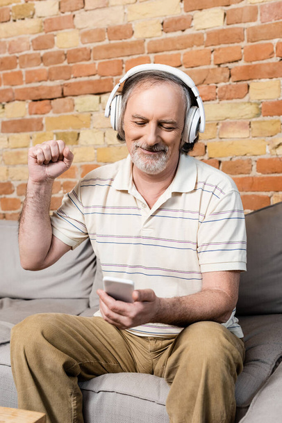 happy and mature man in wireless headphones using smartphone and gesturing in living room  - Foto, afbeelding