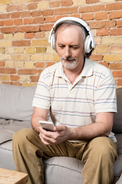 érett férfi vezeték nélküli fejhallgató segítségével okostelefon a nappaliban  - Fotó, kép