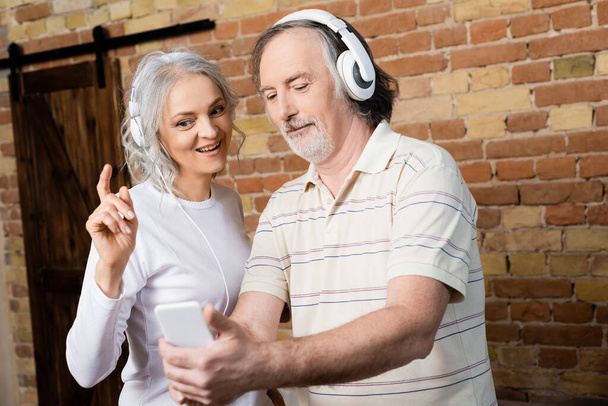 selektywne skupienie wesołej dojrzałej kobiety w słuchawkach bezprzewodowych wskazując palcem na męża ze smartfonem - Zdjęcie, obraz