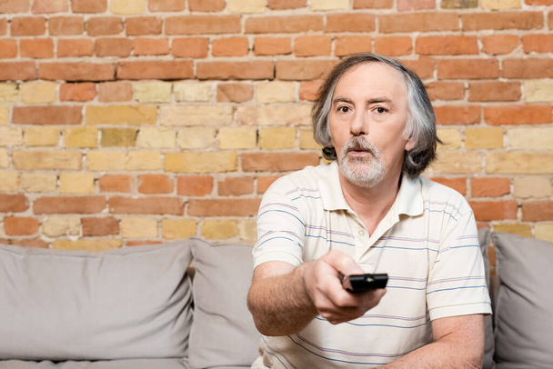 selective focus of surprised and mature man holding remote controller while watching movie at home  - Fotografie, Obrázek