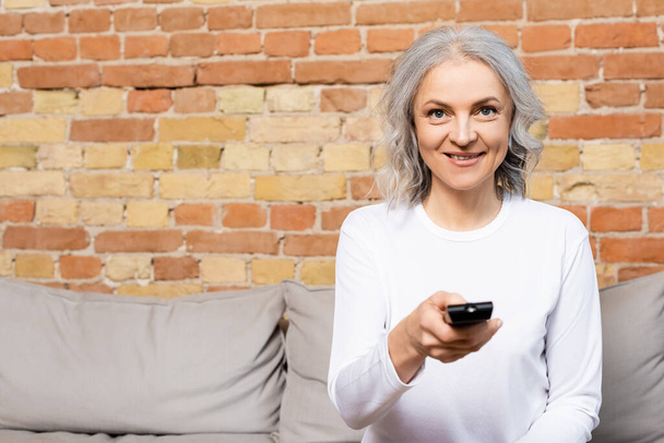 gelukkig en volwassen vrouw holding remote controller tijdens het kijken naar film thuis  - Foto, afbeelding