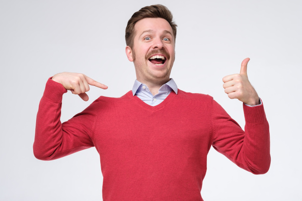 Счастливый бородатый мужчина в красном свитере показывает себя с большими пальцами, улыбается довольный счастливым, указывая на свою грудь, хвастаясь. Я потрясающая. Снимок студии
 - Фото, изображение