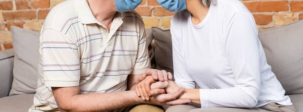 horizontal concept of mature couple in medical masks holding hands while sitting on sofa  - Photo, Image