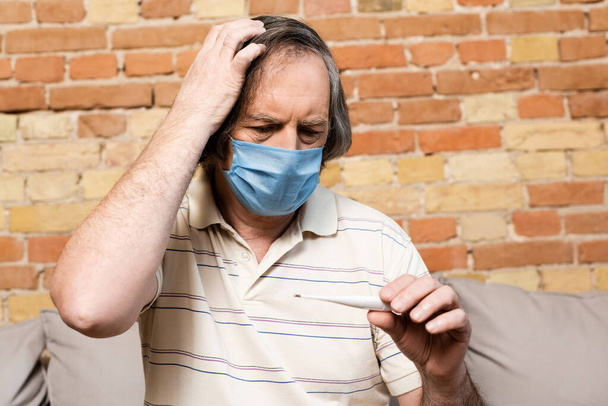 ώριμος άνδρας με ιατρική μάσκα κοιτάζοντας ψηφιακό θερμόμετρο  - Φωτογραφία, εικόνα