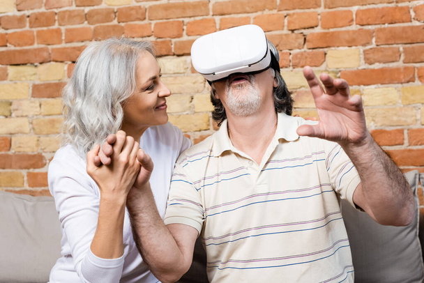ευτυχισμένη και ώριμη γυναίκα κοιτάζοντας τον σύζυγο στην εικονική πραγματικότητα ακουστικά gesturing στο σπίτι  - Φωτογραφία, εικόνα