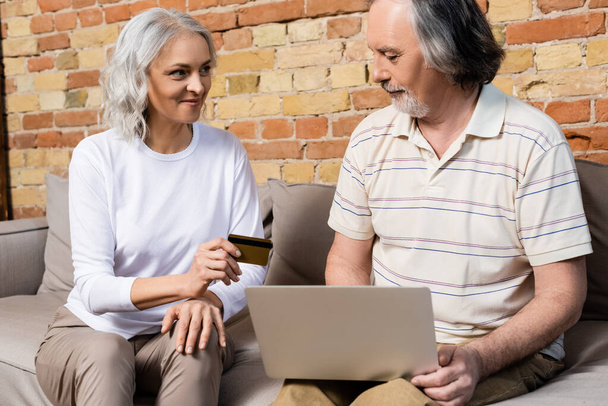 happy and mature woman holding credit card near laptop and middle aged husband  - Photo, Image