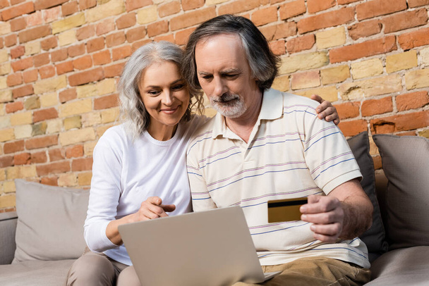 selective focus of mature woman looking at laptop near middle aged husband with credit card  - Fotoğraf, Görsel