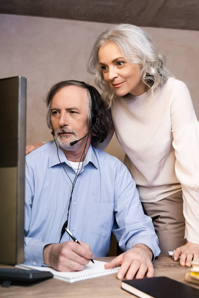 selektywne skupienie operatora w średnim wieku w zestawie słuchawkowym patrząc na monitor komputera w pobliżu żony  - Zdjęcie, obraz