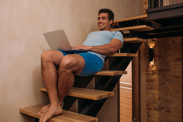 emotional mixed race man using laptop on stairs at home on quarantine - Fotó, kép