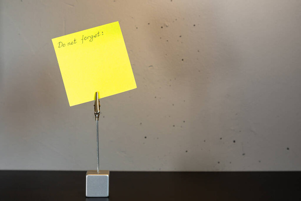 Die Aufschrift "Vergiss nicht" auf einem gelben Blatt Papier, das an einer Klemme befestigt ist, die auf einer schwarzen Fläche an einer grauen Betonwand steht. - Foto, Bild