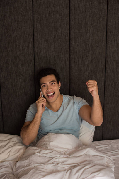 emocionado hombre de raza mixta hablando en el teléfono inteligente en la cama durante el aislamiento
 - Foto, imagen