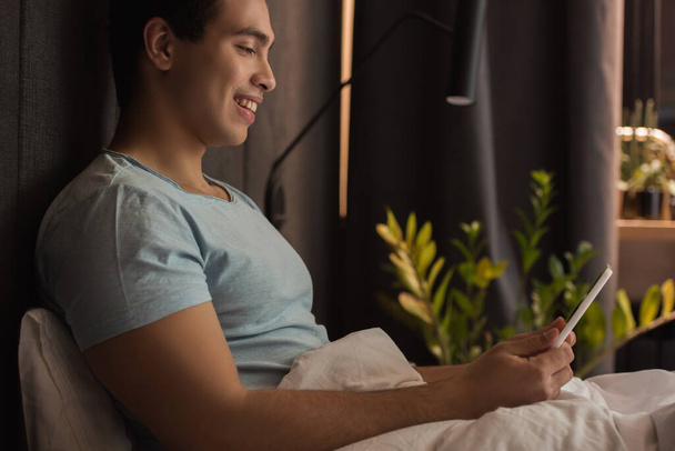 smiling handsome mixed race man using digital tablet in bed during quarantine - Фото, изображение