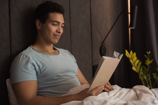 sonriente mestizo hombre leyendo libro en la cama durante auto aislamiento
 - Foto, imagen