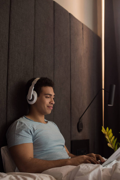 Двурасовый человек слушает музыку с наушниками и ноутбуком в постели на карантине
 - Фото, изображение