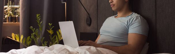 分離中にベッドでノートパソコンでテレワークする混合人種の男のクロップドビューウェブサイトのヘッダー - 写真・画像