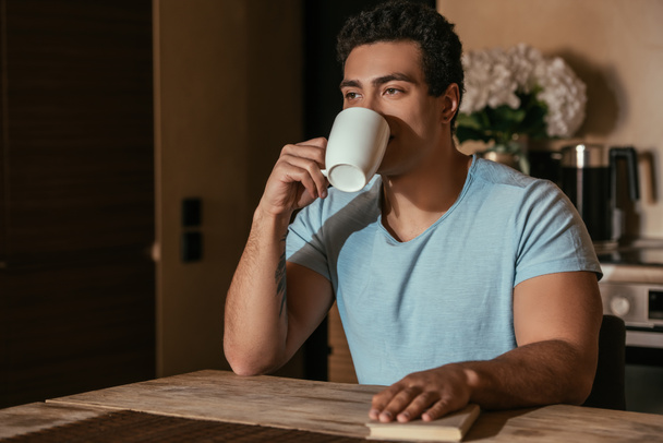 mixed race man drinking coffee and holding book on kitchen during self isolation - 写真・画像