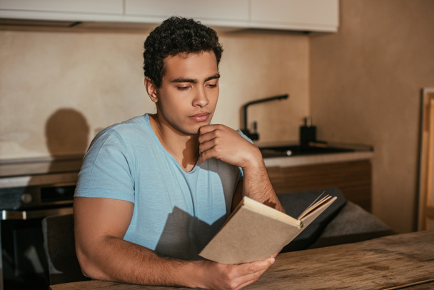 reflexivo hombre bi-racial leer libro en la cocina durante la cuarentena
 - Foto, Imagen