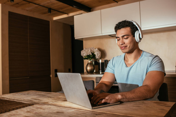 happy mixed race man listening music with headphones and using laptop during self isolation on kitchen - Foto, Imagen