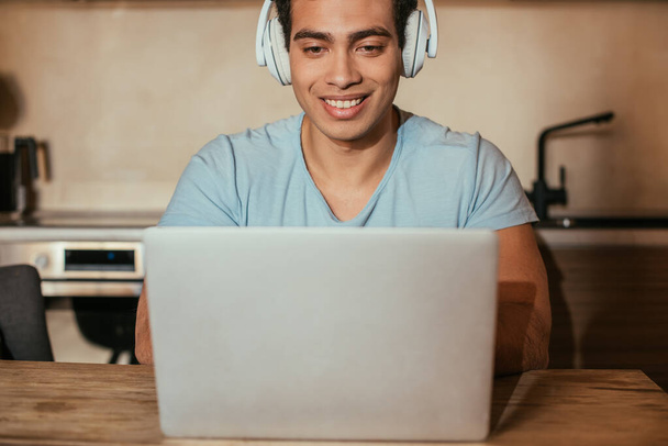 hombre sonriente guapo escuchar música con auriculares y el uso de ordenador portátil durante el autoaislamiento en la cocina
 - Foto, Imagen