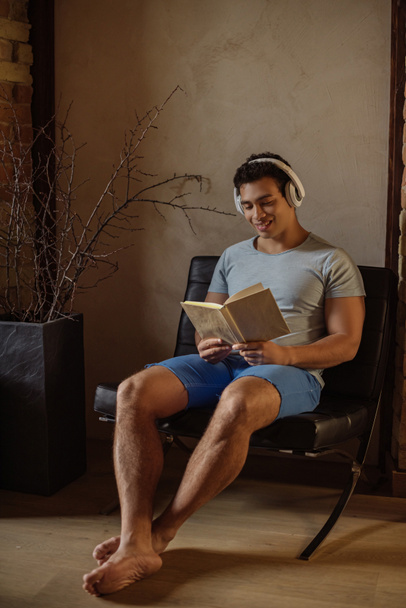 happy mixed race man reading book while listening music with headphones at home during quarantine - Foto, afbeelding